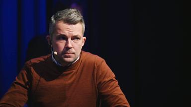 Ein Mann mit kurzen Haaren und im braunen Pullover hat ein Mikro an der Wange und schaut ernst mit gerunzelter Stirn.