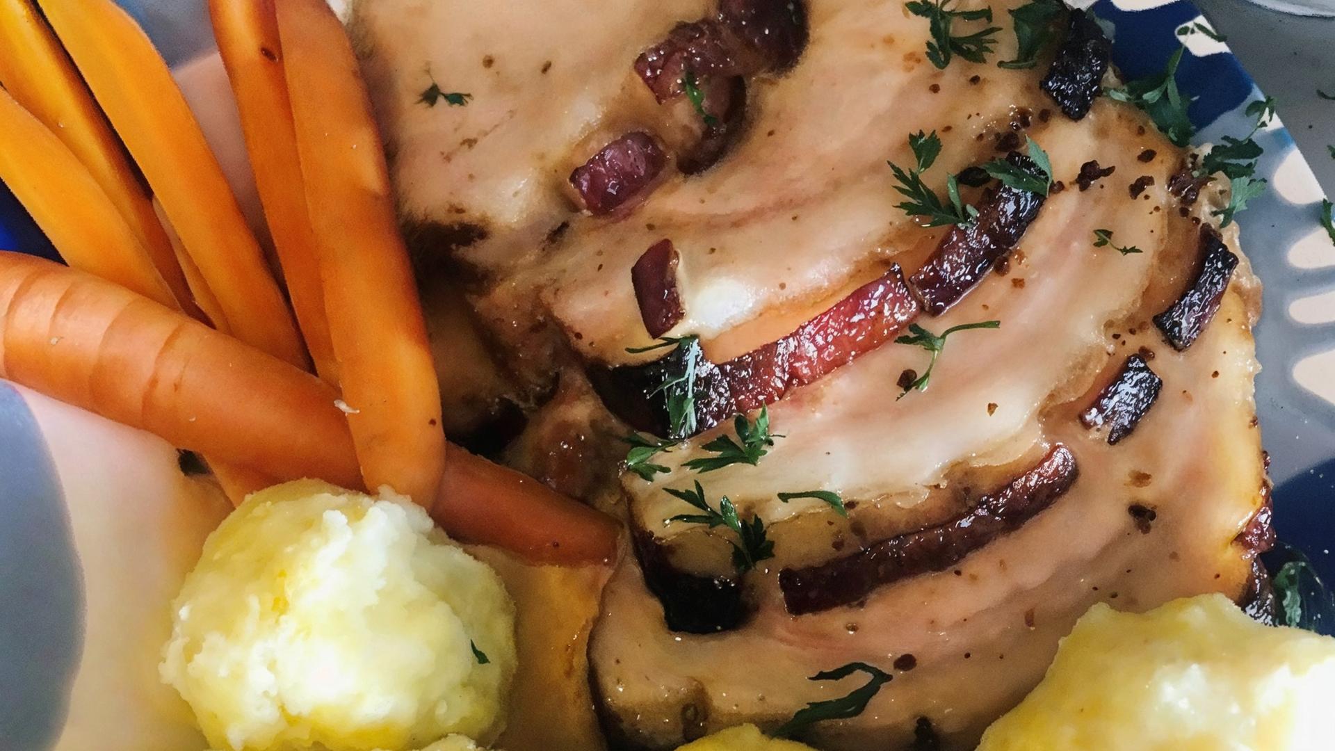 Das Bild zeigt einen Teller mit einem typischen Gericht der österreichischen Küche, insbesondere dem traditionellen "Schweinsbraten". Auf dem Teller liegt eine große, in Scheiben geschnittene Portion Schweinefleisch, das goldbraun und knusprig aussieht. Die Oberfläche des Fleischs ist mit einer dunkleren Kruste versehen und es sind feine Kräuter zur Garnierung sichtbar.  Neben dem Fleisch befinden sich mehrere orangefarbene Karotten, die ungekocht und ungeschält aussehen. Diese sind in länglichen, schmalen Stücken angeordnet.   Auf der anderen Seite des Tellers liegen mehrere knödelähnliche Beilagen, die gelblich und rund sind, wahrscheinlich Mehlknödel. Diese sind weich und haben eine glatte Oberfläche.  Der Teller hat einen farbenfrohen Rand und steht vor einer neutralen Kulisse. Im Hintergrund ist ein kleiner, weißer Schüssel mit frischen Kräutern zu sehen, die als zusätzliche Garnitur dienen könnten.