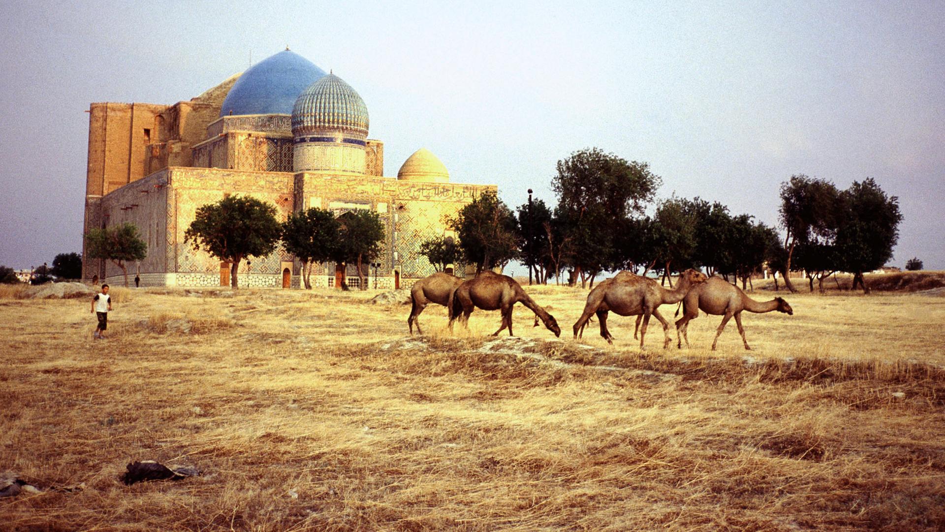 Ein großes Steingebäude in einer Steppe. Im Vordergrund Kamele und ein Kind.