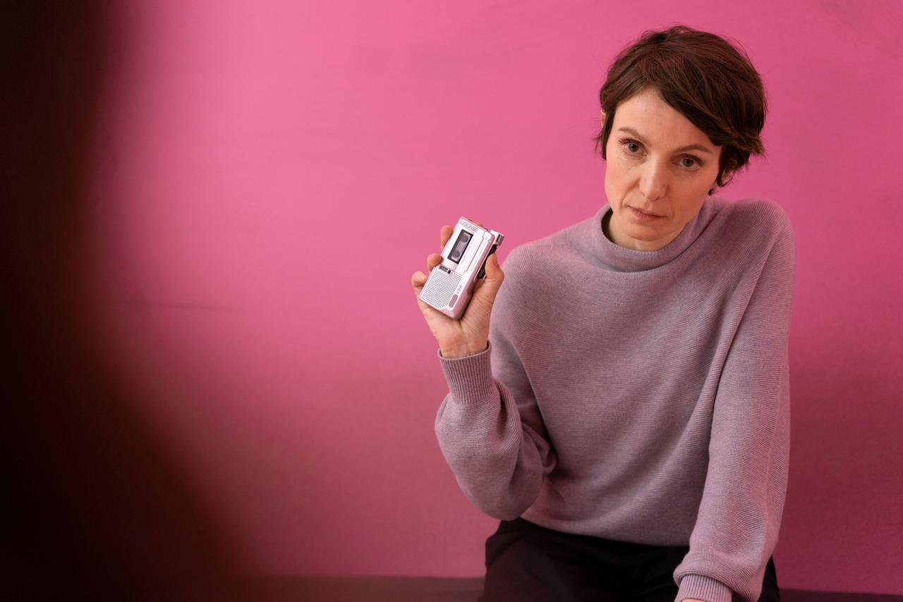 Auf dem Bild sieht man eine Frau mit kurzen, lockigen braunen Haaren, die einen lässigen, grauen Pullover trägt. Sie hält ein tragbares Aufnahmegerät in der Hand. Der Hintergrund ist einfarbig pink und verleiht der Szene eine lebendige Atmosphäre. Der Gesichtsausdruck der Frau ist konzentriert und ernst, während sie in die Kamera schaut. Ihre Pose vermittelt eine gewisse Entschlossenheit, als ob sie bereit wäre, wichtige Informationen zu teilen. Es handelt sich um eine Aufnahme in einem Innenraum.