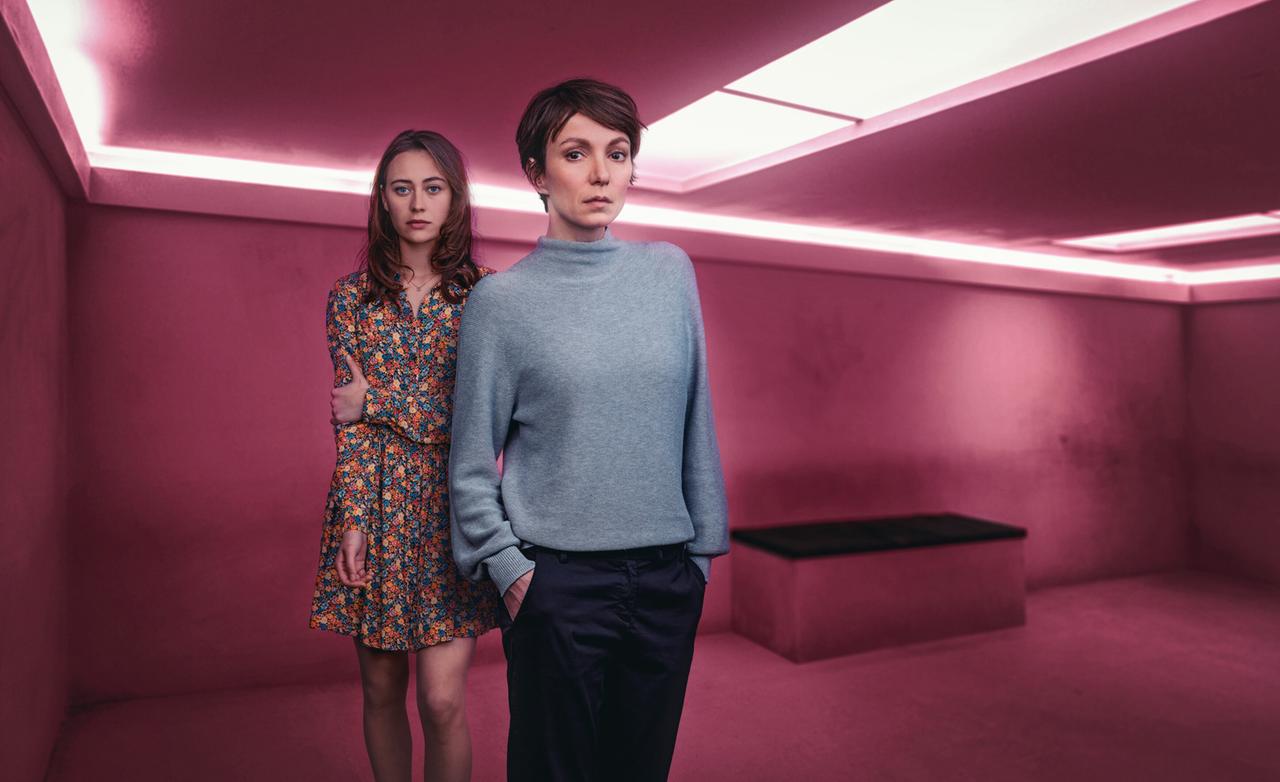 Das Bild zeigt zwei Frauen, die in einem minimalistisch gestalteten Raum mit pinken Wänden und dezentem Licht stehen. Die linke Person trägt ein blumiges Kleid in verschiedenen Farben und hat lockige, mittellange Haare. Sie steht leicht vor der anderen Frau und hat eine nachdenkliche Miene. Die rechte Person trägt einen grauen Pullover und dunkle, lockere Hosen. Sie steht mit einer Hand in der Hosentasche und sieht ebenfalls ernst aus. Im Hintergrund ist eine große, flache Sitzgelegenheit erkennbar. Der Raum hat keine Fenster und wirkt insgesamt sehr modern und steril.