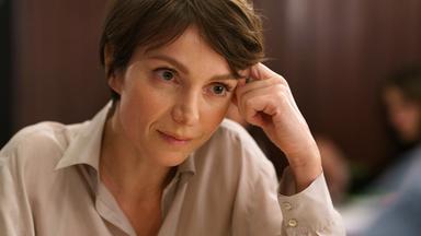 Das Bild zeigt eine Frau mit kurzen, braunen Haaren und einem nachdenklichen Ausdruck. Sie trägt ein helles, offenes Hemd und sitzt an einem Tisch. Ihre linke Hand berührt ihre Schläfe, während sie mit einem intensiven Blick in die Kamera schaut. Der Hintergrund des Bildes ist unscharf, aber man erkennt, dass es sich um einen Innenraum handelt. Es gibt warme, dunkle Holztöne und möglicherweise einige weitere Personen, die unscharf im Hintergrund sitzen. Die Szene vermittelt eine Stimmung der Reflexion oder des Nachdenkens.