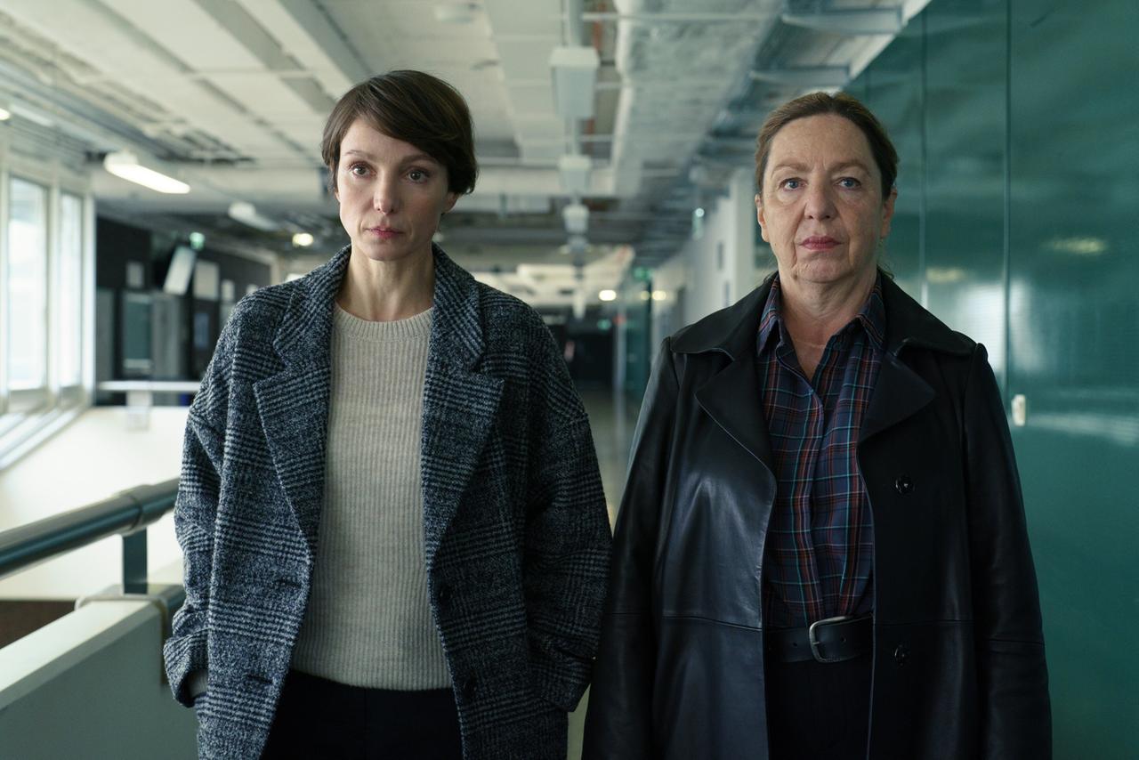 Das Bild zeigt zwei Frauen, die nebeneinander stehen. Die linke Person trägt einen grauen, karierten Mantel über einem hellen Pullover. Sie hat kurzes, braunes Haar und wirkt nachdenklich. Die rechte Person trägt einen schwarzen Ledertrenchcoat sowie ein kariertes Hemd und hat ihre Haare zu einem einfachen Dutt zusammengebunden. Beide schauen ernst und konzentriert in die Kamera.  Im Hintergrund ist ein flurähnlicher Raum mit großen Fenstern zu sehen, durch die natürliches Licht fällt, sowie einer Reihe von Türen und abgehängten Decken mit sichtbaren Rohren und Beleuchtungselementen. Die Wände sind teilweise grün und scheinen eine moderne, büroartige Umgebung zu zeigen.