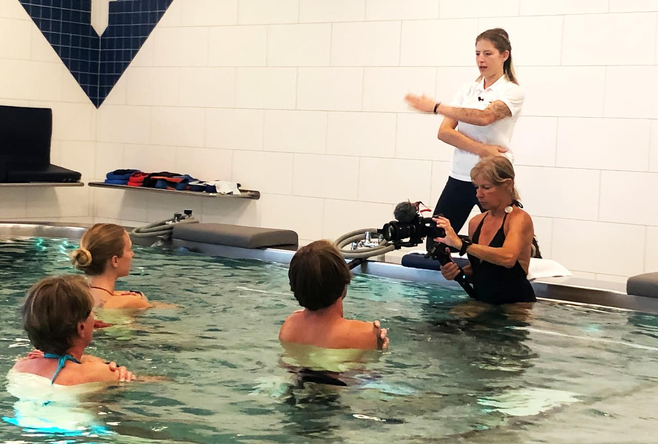 Das Bild zeigt eine Unterwassertherapie in einem modernen Therapiebereich, vermutlich in der Alpentherme Gastein. Im Vordergrund sind vier Personen zu sehen, die im Wasser stehen. Zwei Frauen haben ihre Hände über der Wasseroberfläche, während eine dritte Frau sich mit einem Kameraequipment auf die Gruppe konzentriert. Eine Mitarbeiterin steht am Rand des Therapiebereichs und weist mit den Händen auf Anweisungen hin. Die Wände sind in hellen Fliesen gehalten, und es sind einige Ausstattungsgegenstände wie Sitze und Therapiegeräte sichtbar. Der Raum strahlt eine ruhige und therapeutische Atmosphäre aus.