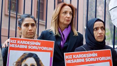 Drei Frauen vor einem Zaun. Zwei der Frauen halten Schilder in der Hand.