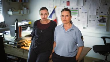 Zwei Frauen stehen nebeneinander in einem Büro vor einem Schreibtisch. Im Hintergrund hängen zahlreiche Dokumente an einer Pinnwand.