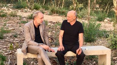 Ferdinand von Schirach und Anselm Kiefer sitzen nebeneinander auf einer Steinbank und unterhalten sich. Beide rauchen.