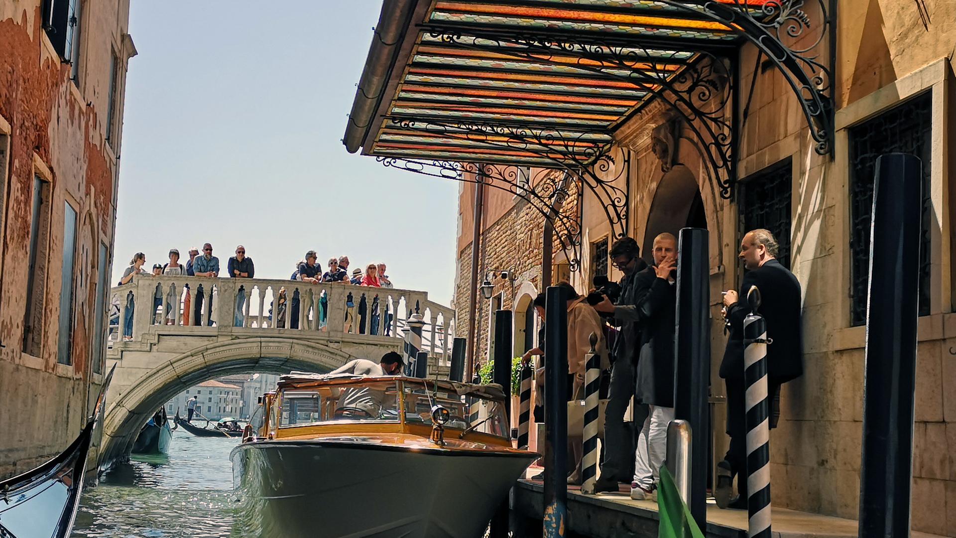 Ein Kanal und eine Brücke in Venedig. Auf der Brücke stehen Menschen. Im Vordergrund ein Motorboot an einem Steeg. Darauf stehen mehrere Personen.