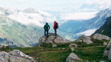 Zwei Wanderer stehen auf einem Felsvorsprung und blicken auf ein weitläufiges, wolkenverhangenes Tal inmitten der Schladminger Tauern; umgeben von felsigem Gelände und grünen Hängen.