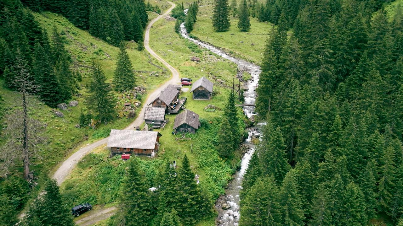 Luftaufnahme einer abgelegenen Berghütte mit mehreren Holzhäusern, einem Bachlauf und umliegendem Waldgebiet inmitten der Schladminger Tauern.