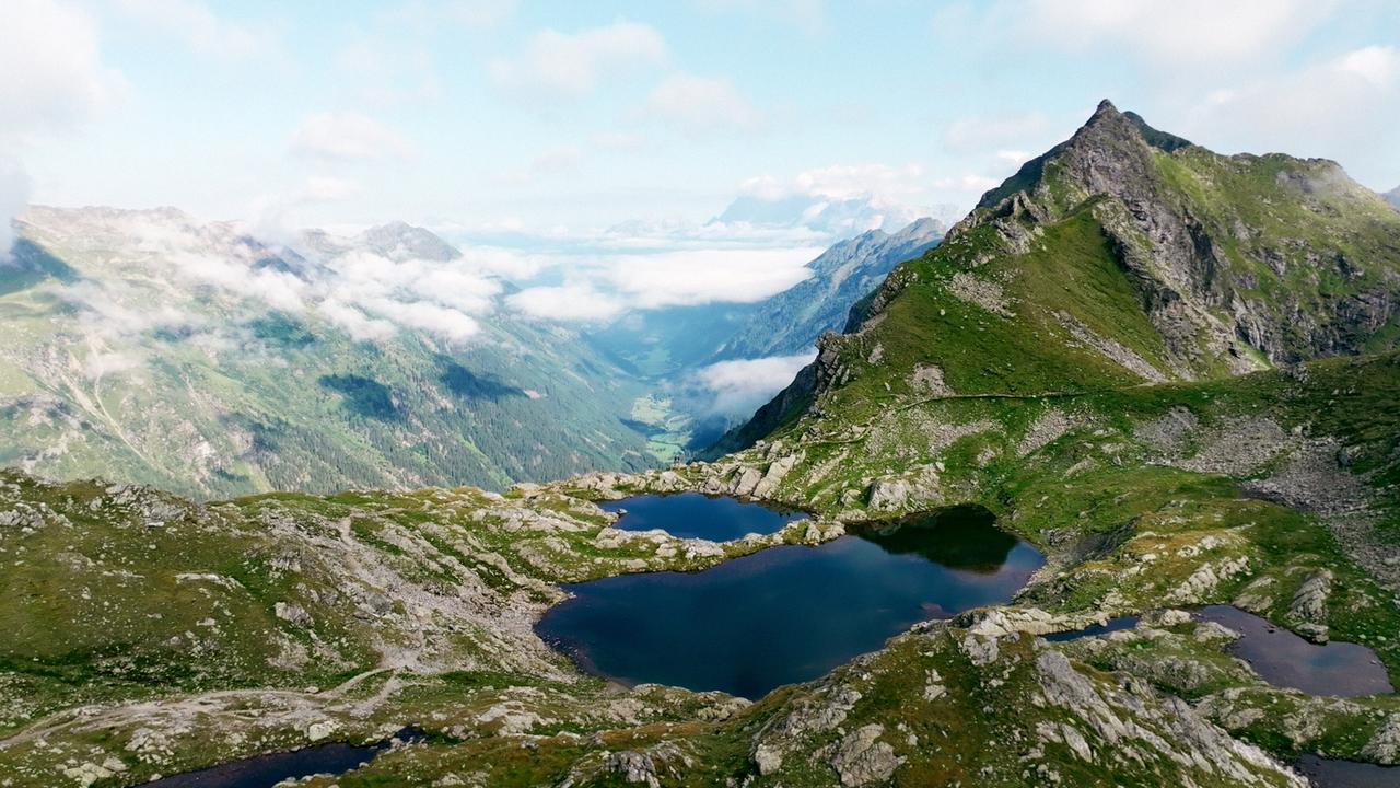 Hochgebirgslandschaft mit mehreren kleinen Seen inmitten grüner Felsen, im Hintergrund ein bewölktes Tal zwischen Berggipfeln der Schladminger Tauern.