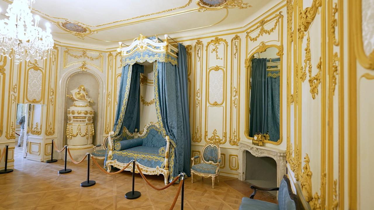 Das Bild zeigt einen Raum im Schloss Esterhazy, einer historischen barocken Residenz. Die Wände sind mit goldenen Verzierungen sowie hellen Tapeten und Dekoren versehen. In der Mitte des Raumes steht ein prächtiges Himmelbett mit blauen Vorhängen und einer reich verzierten Kopf- und Fußseite. Neben dem Bett befindet sich ein eleganter Stuhl mit einer blauen Polsterung. An einer Wand gibt es einen kaminähnlichen Zugang, während eine Statue in einer Nische positioniert ist. Der Boden ist aus Holz und zeigt ein klassisches Muster. An der Decke hängt ein aufwendiger Kristalllüster, und es sind Spiegel und andere dekorative Elemente sichtbar. Geländer aus Seil und Stützen begrenzen den Zugang zum Möbel, um den Raum zu schützen.