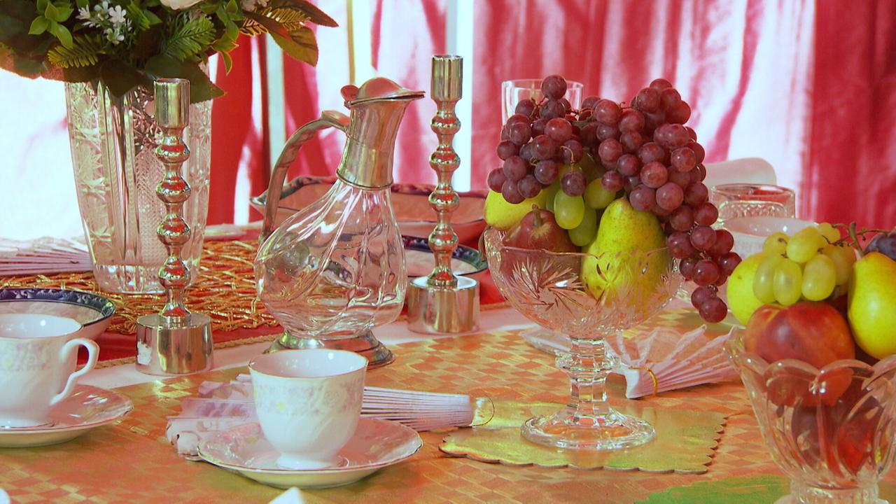 Das Bild zeigt einen festlich gedeckten Tisch in einem historischen Barock-Ambiente. Im Vordergrund befinden sich mehrere feine Teetassen und Unterteller aus Porzellan mit goldenen Verzierungen. Neben den Tassen liegen Servietten, die ordentlich zusammengefaltet sind.

In der Mitte des Tisches steht eine große Glasvase, die mit einer üppigen Anordnung von verschiedenen Früchten gefüllt ist, darunter rote und grüne Trauben, Äpfel und Birnen. Die Glasvase hat einen eleganten Schnitt und steht auf einem Fuß. 

Zwei silberne Kerzenhalter mit einer polierten Oberfläche sind ebenfalls auf dem Tisch platziert, wobei die Kerzenhalter einen dekorativen Look bieten. Zudem gibt es einen klaren Glaskrug, der möglicherweise für Wasser oder eine andere Flüssigkeit gedacht ist.

Die Tischdecke hat ein goldenes Muster und wird im Hintergrund von rötlichen Vorhängen ergänzt, die dem gesamten Setting eine warme, einladende Atmosphäre verleihen. Der Tisch ist sorgfältig dekoriert, was auf ein besonderes Ereignis oder Fest hinweist.