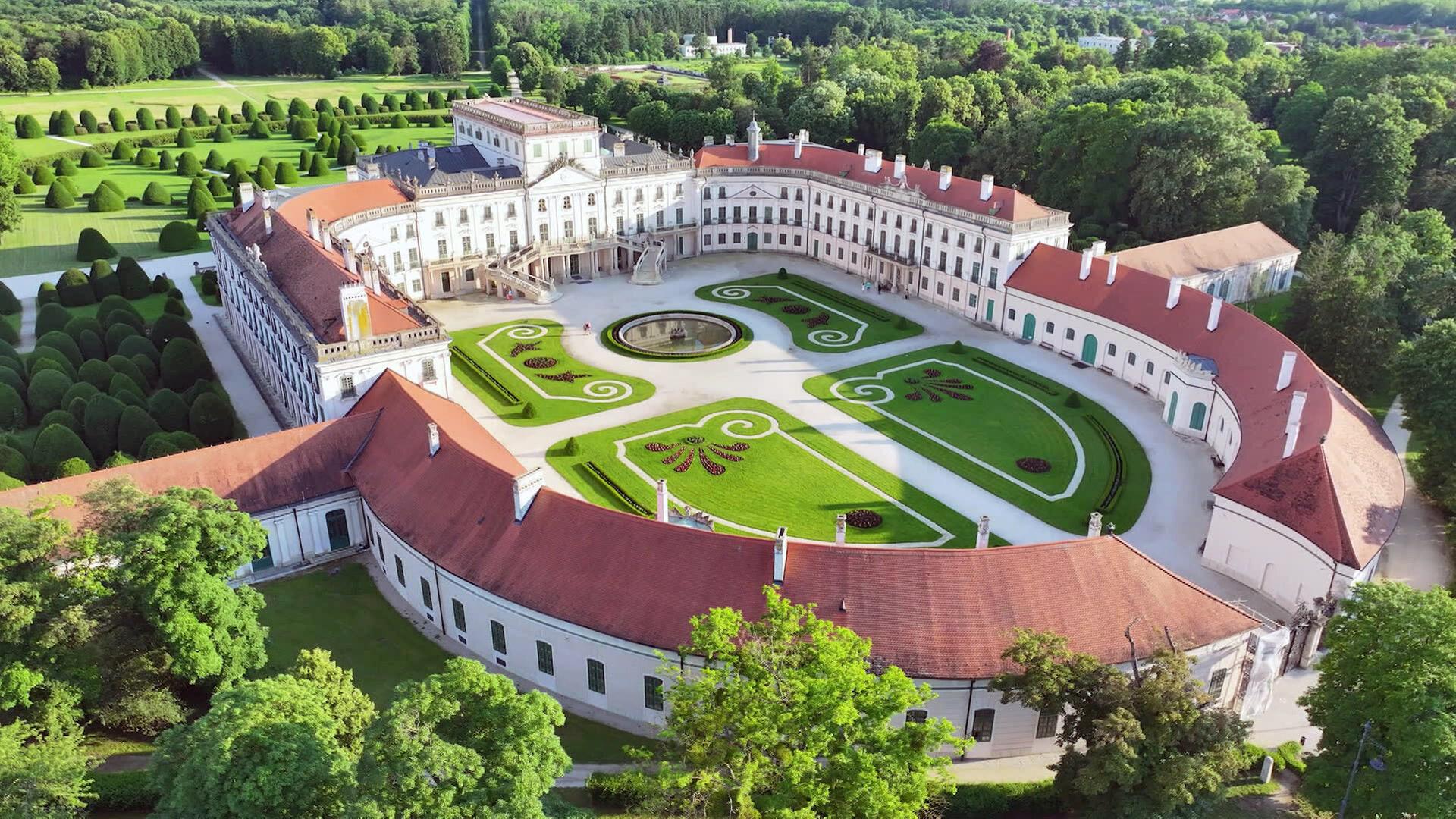 Das Bild zeigt eine Luftaufnahme des Schlosses Esterházy in Fertőd, Ungarn. Im Vordergrund ist ein weitläufiger Schlosspark mit geometrisch angelegten Gärten zu sehen, die mit grünen, runden Hecken und bunten Blumenbeeten geschmückt sind. Das Schloss selbst besteht aus mehreren Gebäudeteilen, die eine elegante, blasse Fassade aufweisen, gekennzeichnet durch große Fenster und mit einem Dach aus roten Ziegeln. In der Mitte des Innenhofs befindet sich ein runder Brunnen, umgeben von geschwungenen Gartenwegen. Im Hintergrund erstreckt sich eine grüne Waldlandschaft. Die Architektur und die Gartenanlagen verleihen dem Ort eine majestätische, barocke Atmosphäre.
