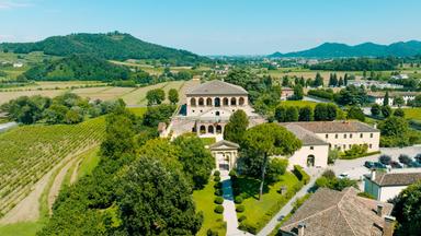 Die Villa Vescovi liegt eingebettet in die Weinlandschaft der Euganeischen Hügel. Das historische Gebäude mit seiner markanten Loggia thront inmitten gepflegter Gartenanlagen, umgeben von sanften Hügeln und grüner Vegetation.