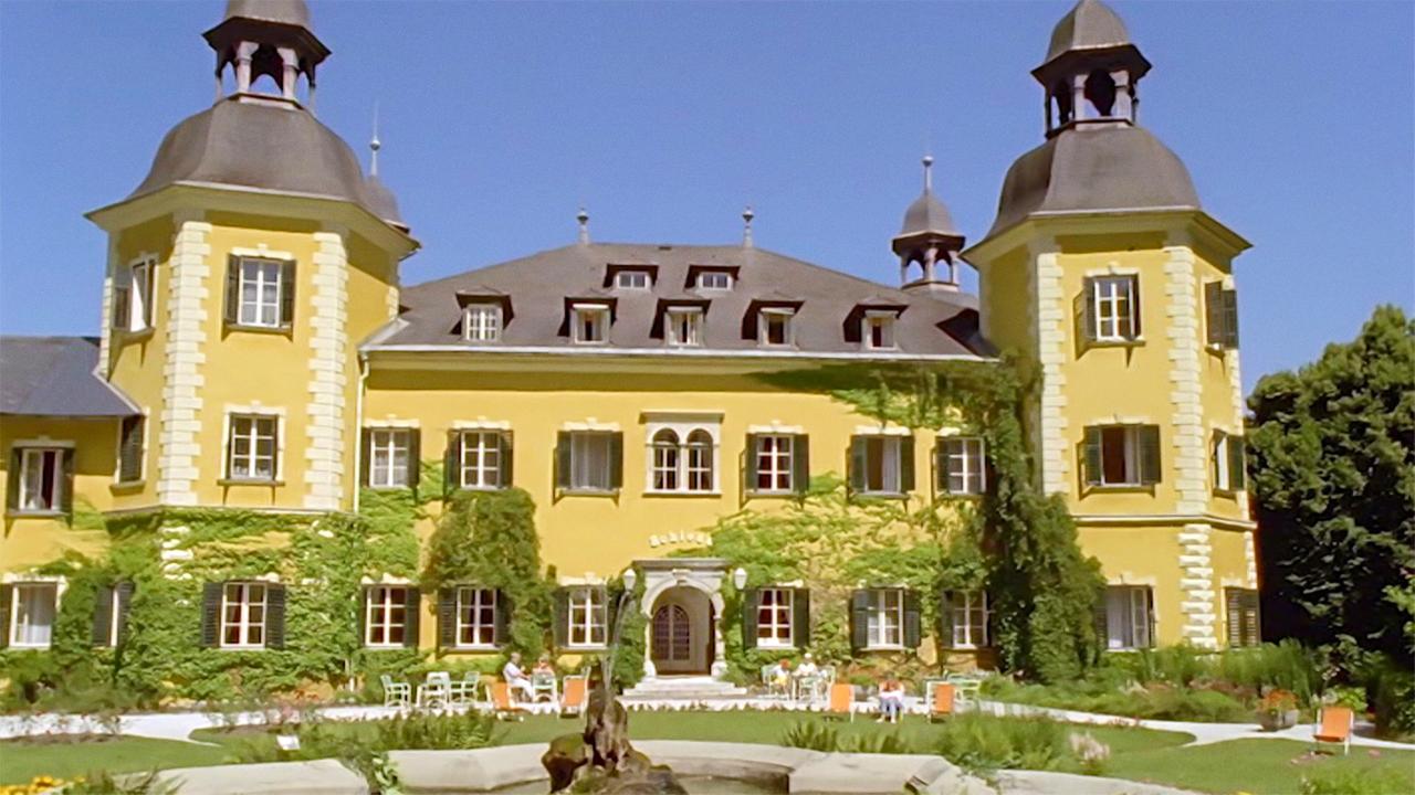 Das Bild zeigt ein historisches Schloss, das als Schlosshotel dient und am Ufer des Wörthersees steht. Die Fassade des Gebäudes ist gelb mit weißen, steinernen Akzenten und wird von Efeu überwuchert. Das Gebäude ist symmetrisch aufgebaut und hat an den Seiten zwei markante Türme mit spitzen Dächern.   Vor dem Schloss ist ein Garten angelegt, in dem mehrere Gäste auf Stühlen sitzen. Diese Stühle sind in Orangetönen gehalten. In der Mitte des Gartens befindet sich ein Brunnen. Der Himmel ist klar und blau, was auf einen sonnigen Tag hinweist.   Diese Szene vermittelt eine Atmosphäre von Erholung und Luxus.