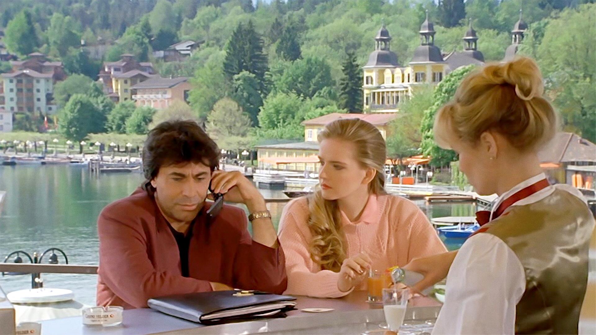 Das Bild zeigt eine Szene aus dem Film "Ein Schloss am Wörthersee". Im Vordergrund befinden sich drei Personen an einem Tisch. Der Mann, der ein rotes Jackett trägt, sitzt am Tisch und hält ein Telefon in der Hand. Neben ihm sitzt eine Frau in einem rosa Pullover, die ihm aufmerksam zuhört. An einem anderen Ende des Tisches steht eine Kellnerin in einer hellen Bluse mit einer schimmernden Weste und serviert ein Getränk.   Im Hintergrund ist eine malerische Seenlandschaft zu sehen, die den Wörthersee darstellt, mit grünen Uferbäumen und mehreren Booten, die am Ufer liegen. Einige bunte Gebäude sind ebenfalls sichtbar, darunter ein großes Gebäude mit Türmchen. Die gesamte Szene vermittelt eine gemütliche, sommerliche Atmosphäre.