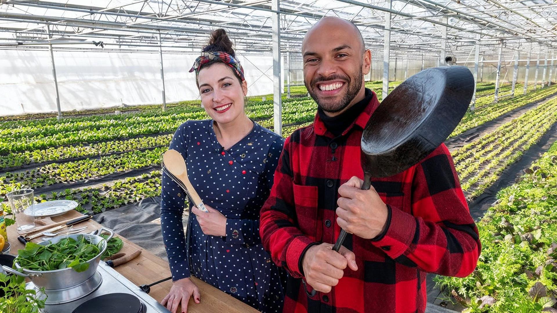 Eine junge Frau und ein junger Mann mit Kochutensilien in einem Gewächshaus.
