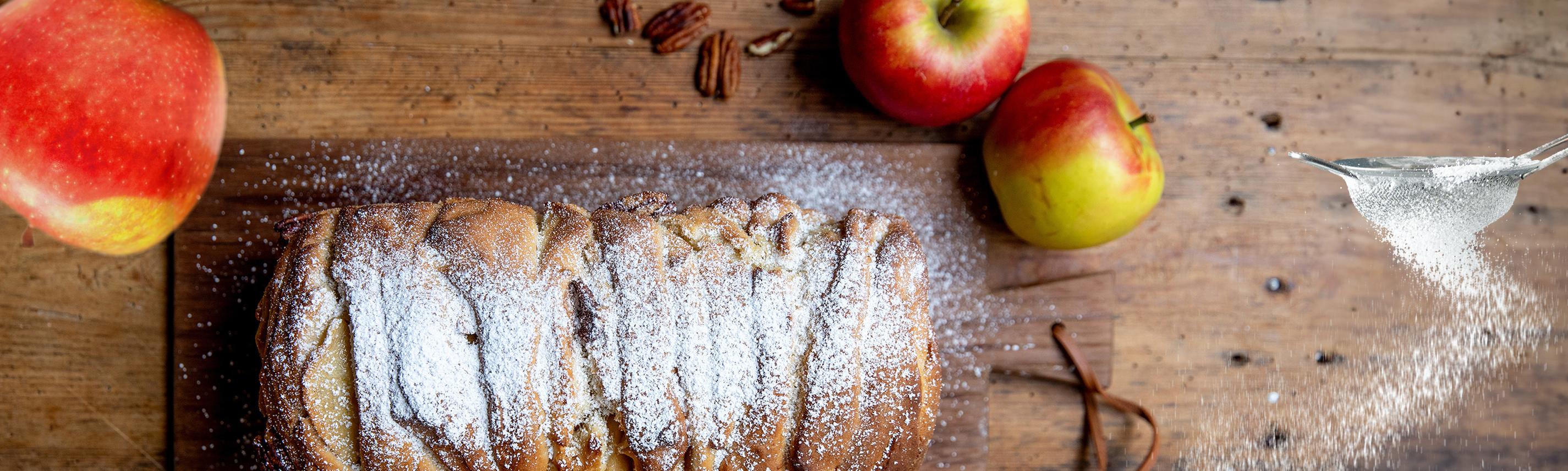 Ein Apfelkuchen, Äpfel und ein Sieb aus dem Puderzucker rieselt, alles auf einem Holzbrett arrangiert