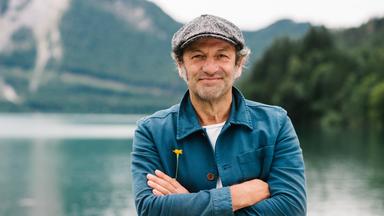 Ein lächelnder Mann mit grauer Schiebermütze und blauer Jacke steht mit verschränkten Armen vor einem Bergsee; an seiner Jacke steckt eine gelbe Blume.