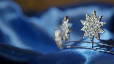 Das Bild zeigt eine Nahaufnahme von zwei funkelnden, sternförmigen Schmuckstücken, die in einem glänzenden blauen Stoff gebettet sind. Die Sterne bestehen aus dem glänzenden Material, das mit vielen kleinen, glitzernden Steinen besetzt ist, was ihnen einen eleganten und opulenten Eindruck verleiht. Der Hintergrund ist weich verschwommen und hebt die Schmuckstücke deutlich hervor. Die Gesamtkomposition vermittelt ein Gefühl von Luxus und Raffinesse, passend zum Thema des Hofjuweliers und der Pracht der Monarchen.