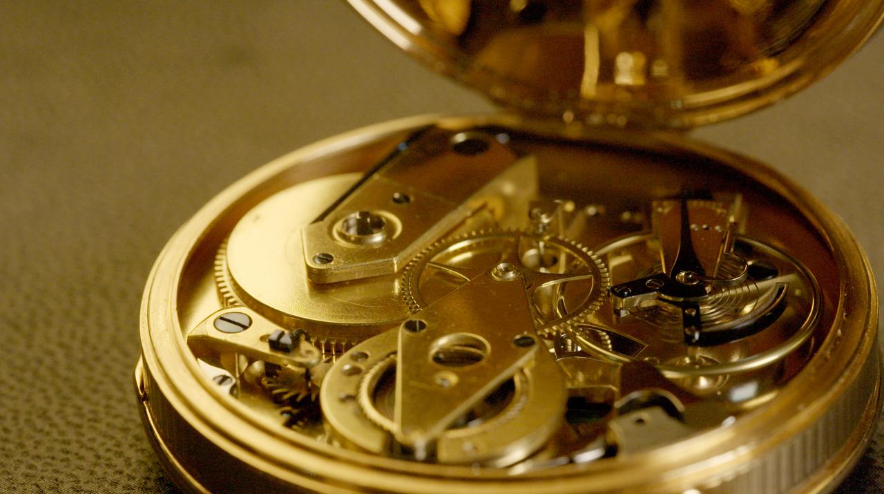 Das Bild zeigt das Innere einer offenen, goldenen Taschenuhr. Der Mechanismus ist gut sichtbar und besteht aus verschiedenen metallischen Komponenten, darunter Zahnräder und Federn, die sich präzise ineinanderfügen. Die goldene Oberfläche der Uhr glänzt, während die dunkleren Teile des Mechanismus, wie Schrauben und einige Federn, einen Kontrast bilden. Der Hintergrund ist neutral und unscharf, was den Fokus auf die Uhr selbst lenkt.   Diese Taschenuhr wird in Verbindung mit dem Thema des Films "Der Schmuck der Monarchen" gebracht, der sich mit der Geschichte und Bedeutung von Hofjuwelen und deren Machteinfluss befasst.