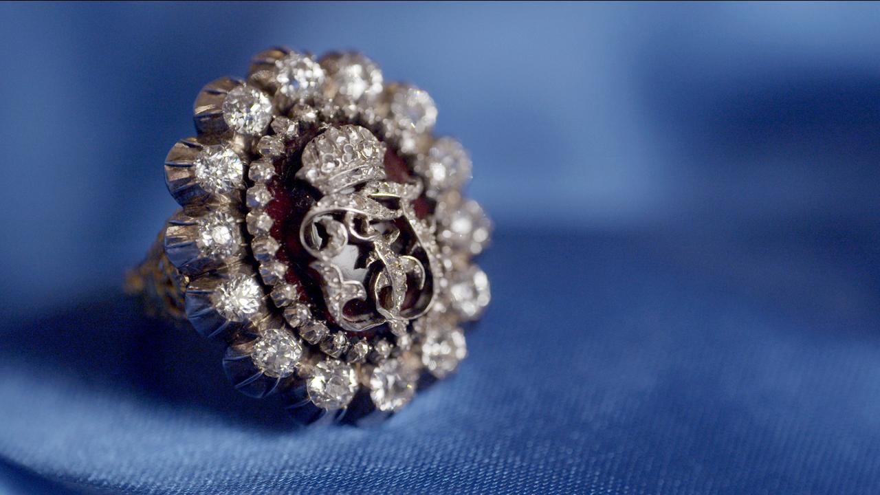 Das Bild zeigt einen prächtigen Ring, der auf einem blauen Stoffhintergrund platziert ist. Der Ring hat eine runde Form und ist von zahlreichen kleinen, funkelnden Brillanten umgeben. In der Mitte des Rings befindet sich ein rotes Element, das den Eindruck eines Monogramms hat, wobei dieses durch eine filigrane, silberne oder metallische Umrandung ergänzt wird. Der Ring strahlt eine opulente und königliche Ausstrahlung aus, was auf seine Zugehörigkeit zu den Monarchen hindeutet. Der Hintergrund ist weich und einfarbig, was den Fokus auf die Details des Rings lenkt.