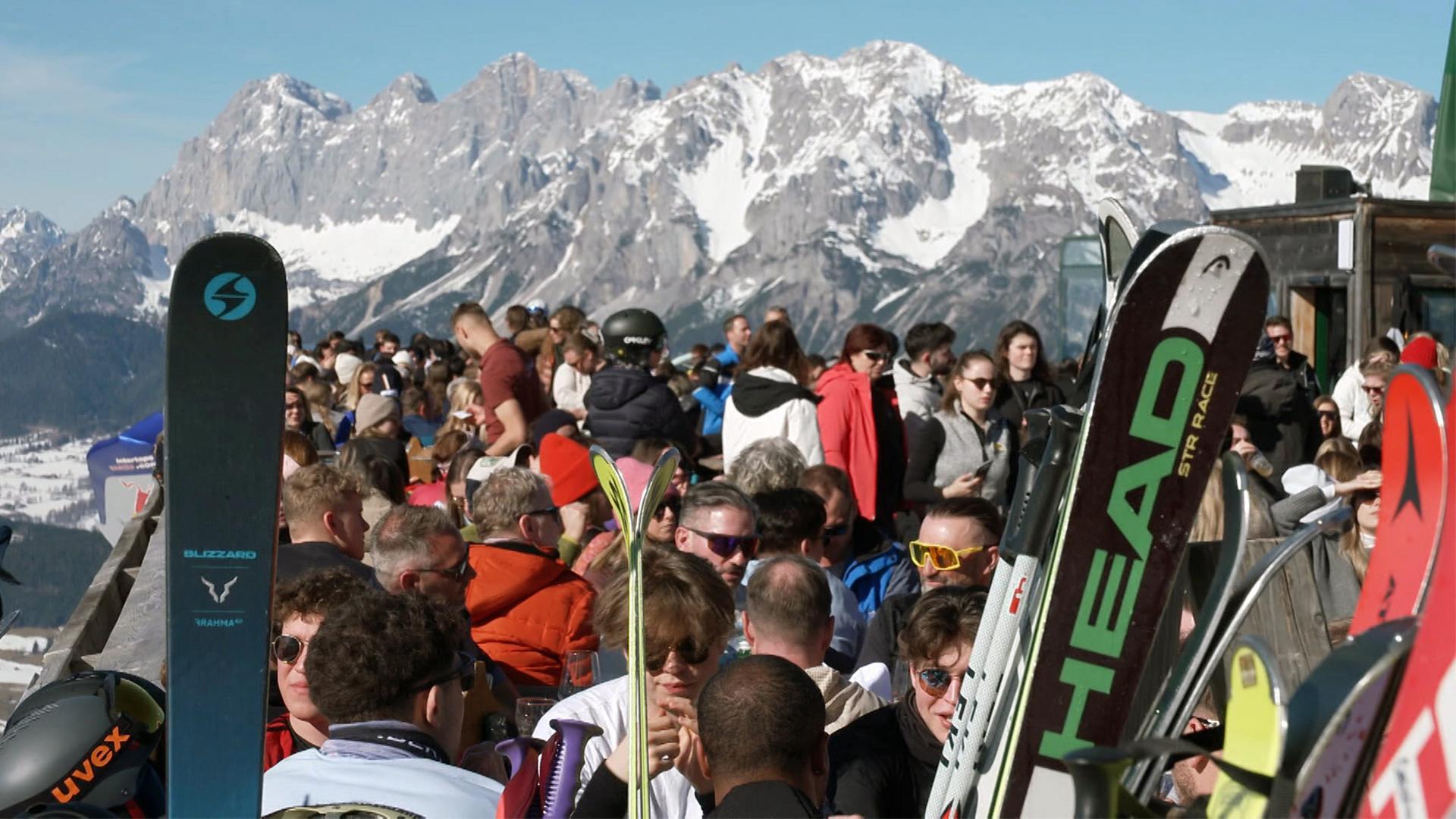 Das Bild zeigt eine große Menschenmenge, die sich in einem Skigebiet in den Alpen versammelt. Im Vordergrund sind mehrere Paar Skier zu sehen, die schräg aufgestellt sind. Die Skier sind mit verschiedenen Markenlogos versehen, darunter „Blizzard“ und „HEAD“.   Die Menschen im Bild stehen dicht beieinander, viele tragen Skibekleidung und einige haben Sonnenbrillen auf. Hintergrund ist eine beeindruckende Berglandschaft mit schneebedeckten Gipfeln, die auf den ersten Blick teilweise bewaldet und an anderen Stellen steinig wirken. Der Himmel ist klar und blau, was auf gutes Wetter hinweist.   Das Bild vermittelt eine lebhafte Szene, die auf die Hochsaison im Skigebiet hinweist, wo laut Beschreibung täglich bis zu 25.000 Menschen anwesend sind. Die Gesamtatmosphäre ist geschäftig und fröhlich, als die Menschen auf den Skitag oder die Freizeitaktivitäten im Schnee warten.