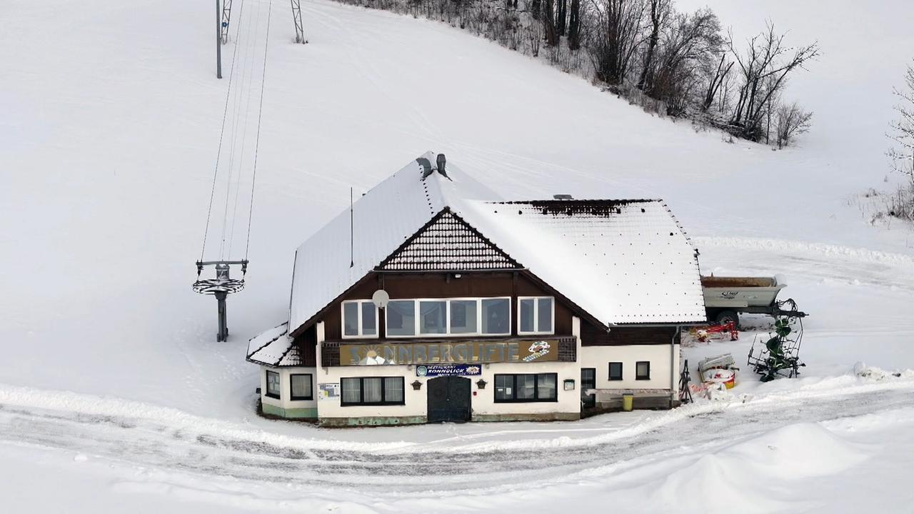 Das Bild zeigt ein heruntergekommenes Gebäude mit dem Schriftzug "Sonnberglifte" an der Fassade. Es befindet sich in einer schneebedeckten Landschaft, umgeben von einer ruhigen, weißen Umgebung. Vor dem Gebäude steht ein leichter Anbau mit Fenstern. Auf der linken Seite ist eine Skiliftanlage sichtbar, die aus einem Gestell mit tragenden Kabeln besteht. Der Boden um das Gebäude ist mit frischem, dickem Schnee bedeckt, und einige Bäume sind im Hintergrund zu erkennen, die teilweise lackiert sind. Die gesamte Szenerie vermittelt einen Eindruck von Einsamkeit und dem Verfall aufgrund der geringen Wintersportaktivität in der Region.