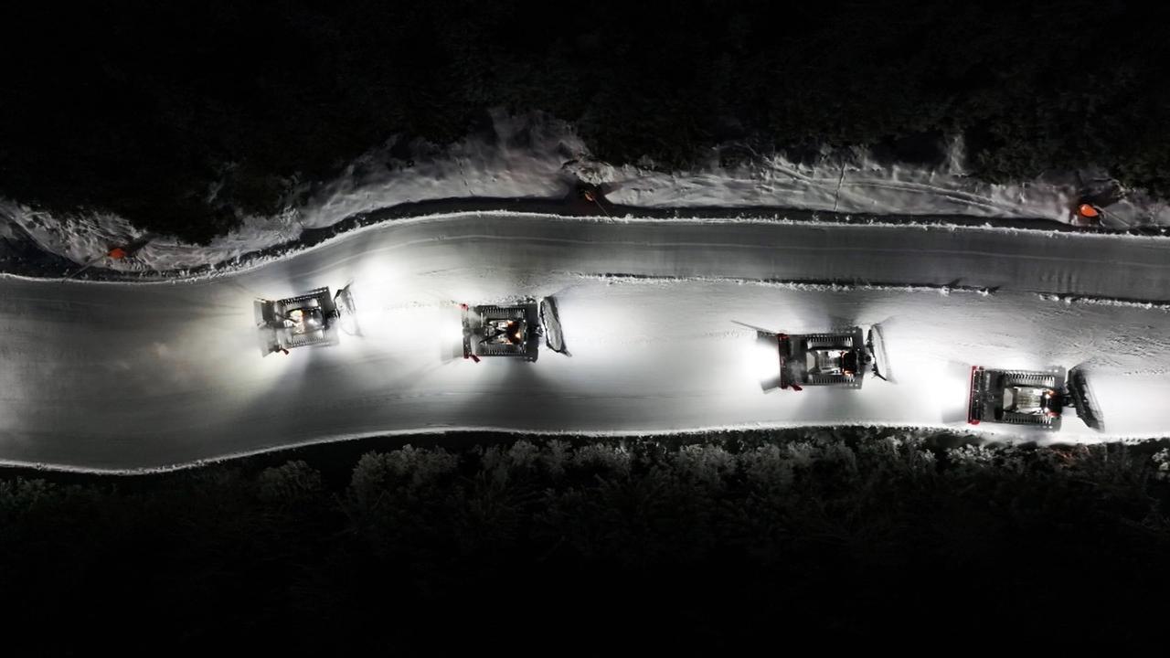Das Bild zeigt eine nächtliche Szene in einem Skigebiet, wahrscheinlich in Schladming, Österreich. Vier Pistenraupen sind auf einer schneebedeckten Skipiste im Einsatz. Sie sind mit starken Scheinwerfern ausgestattet, die den Schnee beleuchten, während sie die Piste präparieren. Die Raupen bewegen sich in einer Linie und hinterlassen eine glatte Schneefläche. Im Hintergrund sind dunkle Baumkronen zu erkennen, die die Szenerie umgeben. Die Atmosphäre ist ruhig und industriell, geprägt von der kontinuierlichen Arbeit, die nötig ist, um die Skipisten für den nächsten Tag vorzubereiten.