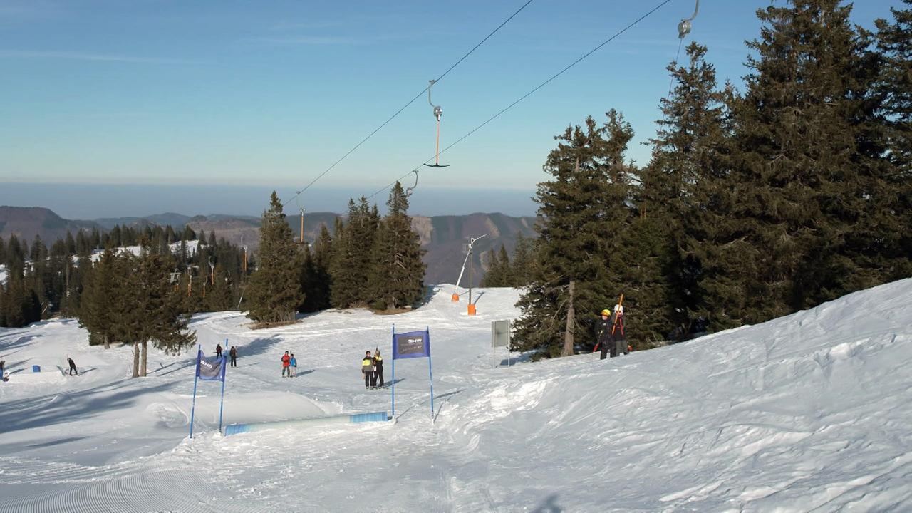 Das Bild zeigt eine Wintersportlandschaft des Familienskigebiets Kasberg in Oberösterreich. Der Vordergrund ist mit frischem, weißem Schnee bedeckt, der teilweise von Skifahrern und Snowboardern genutzt wird. Auf der linken Seite sind ein paar Personen in winterlicher Kleidung sichtbar, die auf Skiern stehen oder sich am Hang bewegen.   Im mittleren Bereich sind blaue Flaggen aufgestellt, die den Verlauf einer Skipiste markieren. Im Hintergrund sind hohe Tannenbäume, die eine natürliche Grenze bilden, sowie einige Skilifte, die im oberen Bereich des Bildes erkennbar sind. Der Himmel ist klar und blau, was auf gute Wetterbedingungen für den Wintersport hindeutet.   Die Szenerie vermittelt eine ruhige, winterliche Atmosphäre, die durch die schneebedeckte Landschaft und die sporadische Anwesenheit von Menschen belebt wird.