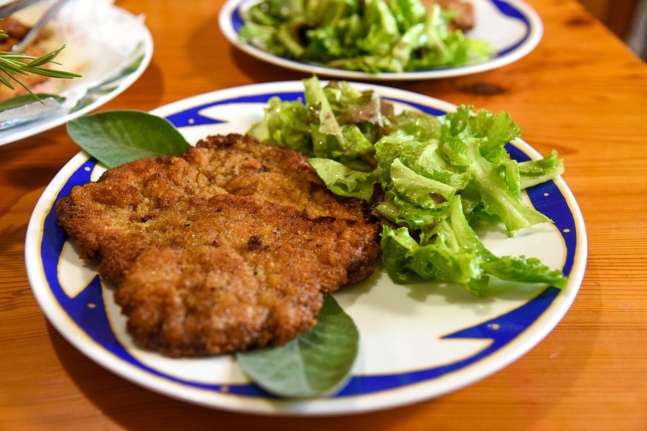 Das Bild zeigt einen Teller mit einer Cotoletta alla Milanese, einem italienischen Schnitzel. Das Schnitzel ist goldbraun und knusprig, auf einem blauen Teller mit einem einfachen, aber dekorativen Rand arrangiert. Neben dem Schnitzel liegt ein frischer Salat mit grünen Blättern, der ebenfalls auf dem Teller serviert ist. Im Hintergrund sind weitere Teller mit Salat und anderen Speisen sichtbar. Der Tisch, auf dem die Teller stehen, ist aus Holz und hat eine warme, natürliche Farbe.