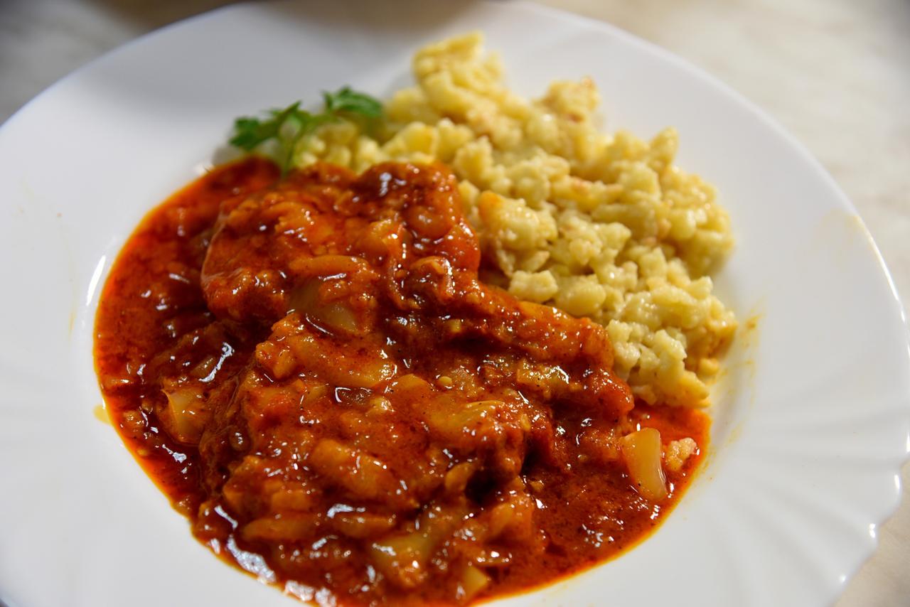 Das Bild zeigt einen Teller mit einem Gericht, das aus zwei Hauptbestandteilen besteht. Links sind Stücke von Fleisch zu sehen, die in einer dicken, roten Sauce serviert werden. Die Sauce hat eine glatte Konsistenz und eine würzige Farbe, und es sind einige fleischliche Zutaten sichtbar.   Rechts vom Fleisch befindet sich eine Portion von Tarhonya, kleinen, genussfertigen Teigwaren, die hellgelb bis beige gefärbt sind. Diese sind weich und haben eine unregelmäßige Form. Der Teller, auf dem die Speisen angerichtet sind, hat einen klassischen, weißen Rand, der das Essen hervorhebt.   Das gesamte Gericht wirkt frisch zubereitet und scheint sehr reichhaltig und schmackhaft zu sein. Hintergrund ist unscharf, sodass der Fokus klar auf dem Essen liegt.