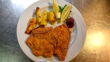 Das Bild zeigt einen Teller mit einem Cordon bleu, das mit einer Schüttelbrotpanade zubereitet ist. Das Cordon bleu ist gefüllt mit Speck und Ziegenkäse. Auf dem Teller befinden sich außerdem einige Stücke gebratene Kartoffeln in unterschiedlichen Formen, die goldbraun angeröstet sind. Dazu sind zwei Zitronenschnitze sowie ein Klecks Preiselbeermarmelade dekorativ angerichtet. Der Teller ist weiß und steht auf einer glänzenden, metallischen Unterlage, die silberfarben aussieht.