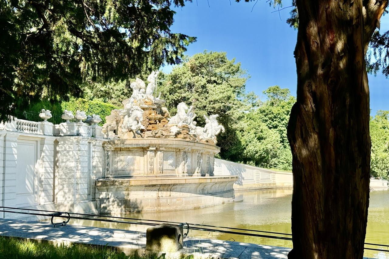 Das Bild zeigt den Schlossgarten von Schloss Schönbrunn in Wien, einem historischen Ort und UNESCO-Weltkulturerbe. Im Vordergrund ist eine große, runde Fontäne zu sehen, die mit kunstvollen Skulpturen verziert ist. Diese Skulpturen stellen mythologische Figuren und Tiere dar und sind in einem hellen Stein gehalten.   Um die Fontäne herum befindet sich ein ruhiger Wasserspiegel. Im Hintergrund sind grüne Bäume und Büsche zu erkennen, die eine natürliche Kulisse bieten. Es gibt eine klare, blaue Himmelssicht, die das Licht über die gesamte Szene verteilt. An den Rändern des Bildes sind teilweise Schatten von Bäumen zu sehen, was einen Kontrast zu den helleren Bereichen schafft.   Diese Darstellung spiegelt die barocke Architektur und die botanische Vielfalt des Gartens wider, die einst ein kaiserliches Jagdrevier waren und heute als Ort für kulturelle Ereignisse und Erholung dienen.