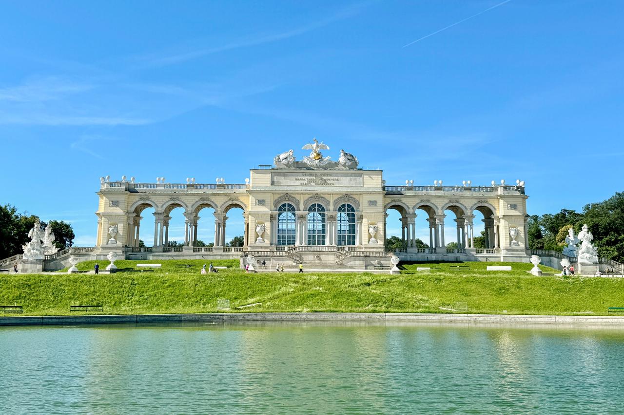 Das Bild zeigt die Gloriette im Schlossgarten von Schloss Schönbrunn in Wien. Die Gloriette ist ein beeindruckendes Bauwerk im barocken Stil, das auf einem erhöhten Platz steht und von einer gepflasterten Fläche umgeben ist. Sie hat große, offene Bögen und Fenster, die viel Licht hereinlassen. Auf dem Dach der Gloriette befindet sich eine Statue, die einen Adler darstellt.  Vor der Gloriette erstreckt sich ein grüner Rasen, der sanft zum Wasser eines Teiches hin abfällt. Auf der Wiese sind mehrere Menschen zu sehen, die umhergehen und das Gelände erkunden. An den Seiten der Gloriette stehen monumentale Skulpturen, die das Gesamtbild ergänzen. Im Hintergrund sind einige Bäume sichtbar.  Der Himmel ist klar und blau, was dem Bild eine helle und freundliche Atmosphäre verleiht. Die Gloriette ist ein symbolträchtiger Ort innerhalb des Schlossgartens, der als Teil des UNESCO-Weltkulturerbes gilt.