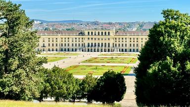 Auf dem Bild ist das Schloss Schönbrunn zu sehen, ein imposantes historisches Gebäude in Wien, Österreich. Das Schloss hat eine helle, gelbe Fassade mit mehreren großen Fenstern und einem symmetrischen Design. Vor dem Schloss erstreckt sich ein großer Garten, in dem geometrisch angelegte Blumenbeete und Rasenflächen sichtbar sind. Die Beete sind in verschiedenen Farben gestaltet und zeigen eine ordentliche, festliche Anordnung.   Im Vordergrund des Bildes sieht man Bäume, die den Garten an den Seiten einrahmen, und es gibt eine hügelige Wiese im Vordergrund. Auf dem Platz vor dem Schloss sind einige Besucher zu sehen, die spazieren gehen. Der Himmel ist klar und blau, mit nur wenigen kleinen Wolken. Die Landschaft um das Schloss herum zeigt die Stadt Wien im Hintergrund, mit Gebäuden und Hügeln.   Insgesamt vermittelt das Bild den Eindruck eines prächtigen, kulturell bedeutsamen Ortes, der sowohl historische als auch botanische Schönheit bietet.