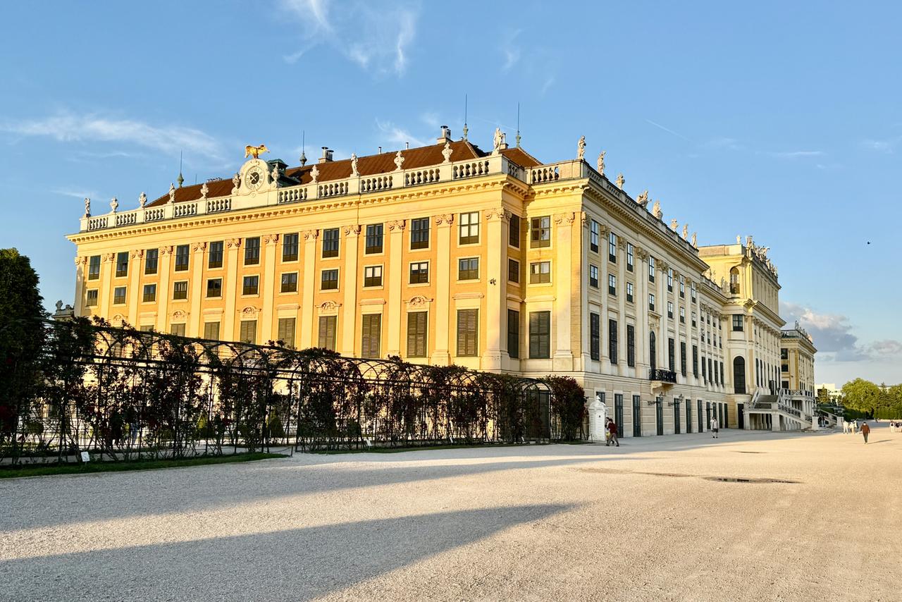 Das Bild zeigt das Schloss Schönbrunn in Wien, ein historisches Gebäude, das sowohl als kaiserliche Residenz als auch als Gartenanlage bekannt ist. Die Architektur ist barock geprägt, mit einer gelben Fassade, großen Fenstern und einem Dach, das mit Skulpturen und Ornamenten verziert ist. Über dem Hauptgebäude befindet sich eine goldene Figur.  Im Vordergrund sieht man einen weitläufigen Gartenbereich mit einem Kiesweg, der vom Schloss wegführt. Auf der linken Seite des Bildes sind kunstvoll gestaltete Gartenanlagen zu erkennen, die von einer Gitterstruktur umrahmt werden, in der Pflanzen wachsen.   Der Himmel ist blau und es sind einige Wolken sichtbar, was auf eine klare Tageszeit hindeutet. Die Lichtverhältnisse deuten auf den späten Nachmittag oder Abend hin, da das Schloss in warmes Licht getaucht ist. Es ist eine ruhige, elegante Atmosphäre zu spüren, die den historischen Charakter des Ortes unterstreicht.