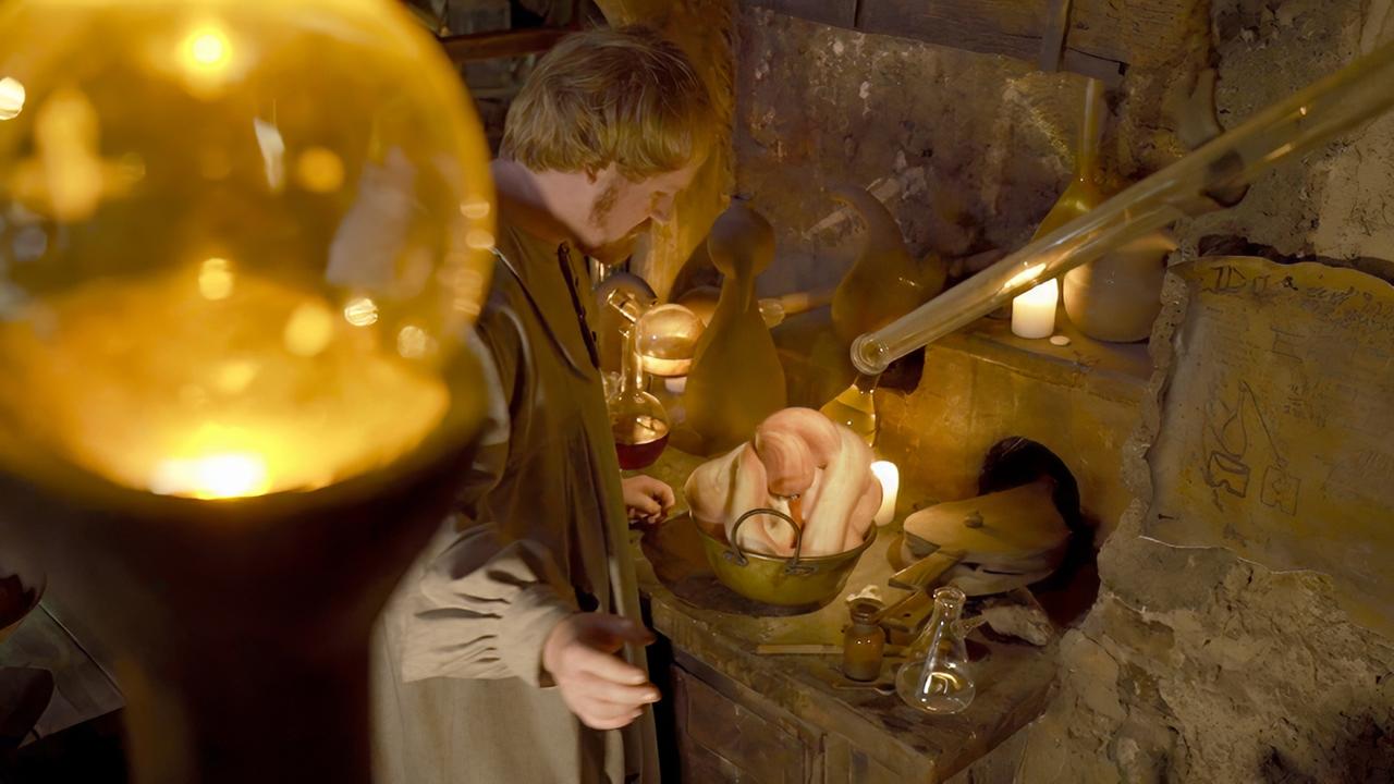 Ein Mann in historischer Kleidung steht in einem nachgebauten alchemistischen Labor, das mit Glasgefäßen, Retorten und Kerzen beleuchtet ist. In einem Kessel liegt eine geheimnisvolle Figur, die aussieht wie ein künstlich erzeugtes Wesen.