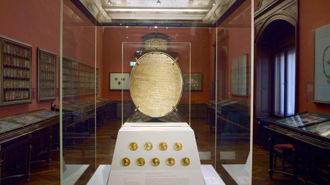 In einem prunkvollen Museumsraum ist eine goldene Gedenkmedaille mit lateinischer Inschrift in einer Glasvitrine ausgestellt. Darunter sind mehrere goldene Münzen mit Porträts zu sehen. Die Wände sind mit weiteren Medaillen und Orden dekoriert.