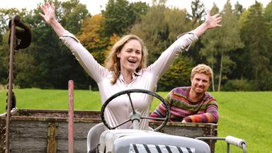 Eine Frau am Steuer eines Traktors hat die Hände in die Luft gestreckt. Hinter ihr sitzt ein Mann.