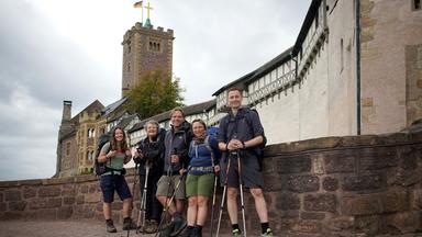 "Schritt für Schritt in die Freiheit": Drei Frauen und zwei Männer stehen mit Outdoorkleidung, Walking-Stöcken und Rucksäcken vor einem Gemäuer, im Hintergrund eine Burgfestung. Sie schauen alle lächelnd in die Kamera