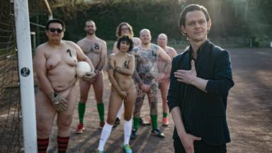 Mann in Anzug in Torwartschützpose mit gemischten nackten Fussballteam im Tor hinter ihm