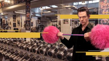 Moderator Jo Schück im  Fitnessstudio, umgeben von aufgereihten Kurzhanteln aus Metall. Er stemmt eine Hantel mit pinken Plüschgewichten. 