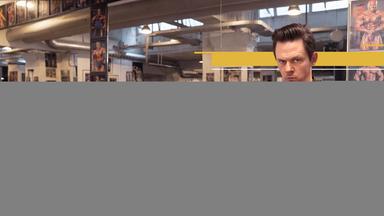 Moderator Jo Schück im  Fitnessstudio, umgeben von aufgereihten Kurzhanteln aus Metall. Er stemmt eine Hantel mit pinken Plüschgewichten. 