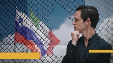 Jo Schück starrt nachdenklich auf den Grenzzaun von Nova Gorica, hinter dem eine Italienische und eine slowenische Flagge gehisst sind.