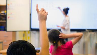 Szene in einer Schulklasse: Ein Kind hebt die Hand, um sich zu melden, während die Lehrerin an die Tafel schreibt.