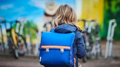 Schülerin von hinten mit Schulranzen auf dem Rücken aufr dem Weg in die Schule.