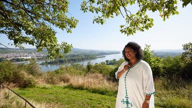 Menna Mulugeta steht vor einem Landschaftspanorama des Rheins. Sie fasst mit einer Hand an einen Kettenanhänger in Form von Afrika.