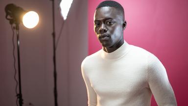 Alpha Dia, ein junger, Schwarzer Mann in weißem Pullover, steht vor einer pinkfarbenen Wand. Neben ihm stehen zwei Stehlampen.